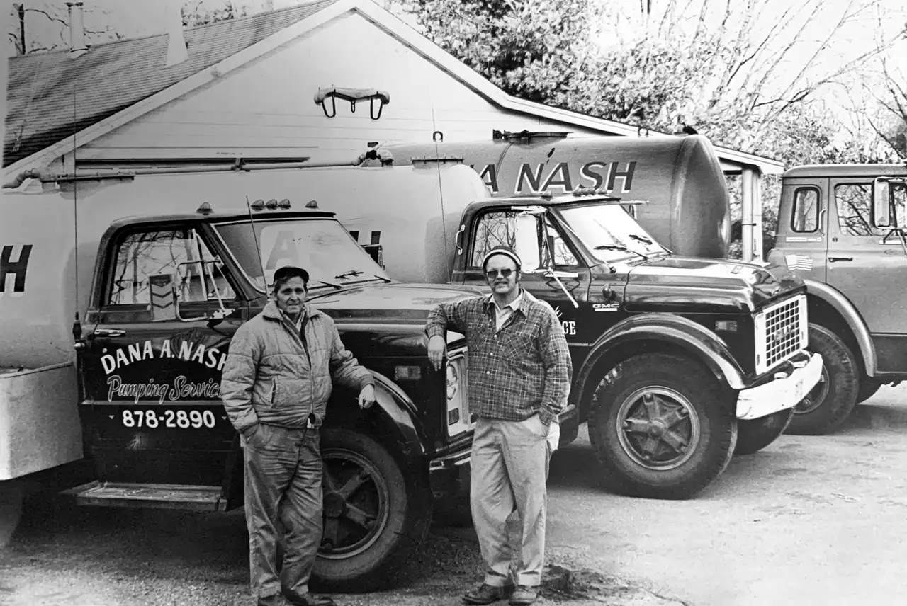 Black-and-white photo of Dana A. Nash Jr. (right) and a worker standing by Dana A. Nash Pumping Service tank trucks outside the family garage in Abington