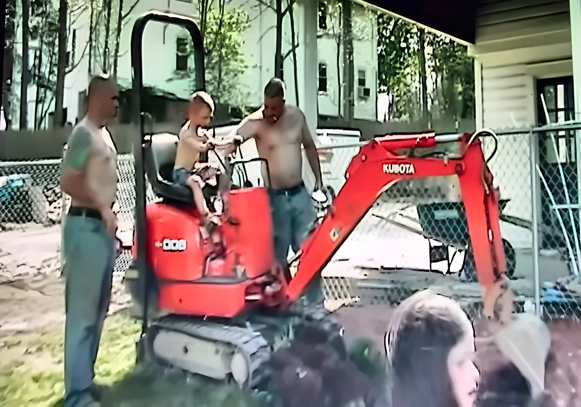 Johnny as a little boy in the seat of a red Kubota mini excavator, with his dad standing on the left and his uncle Tony on the right