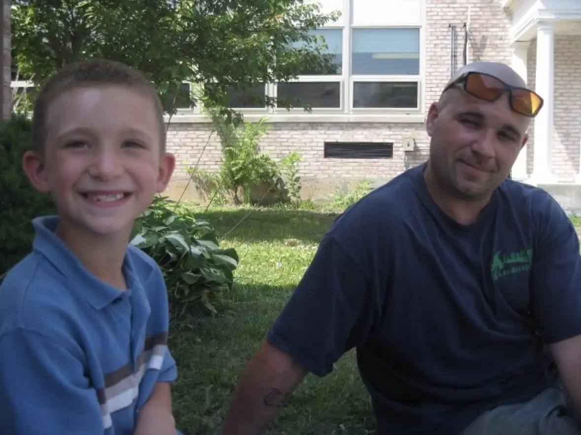 Johnny as a young boy, smiling, sitting on the grass beside his dad, who wears a J DeMarco Landscaping shirt
